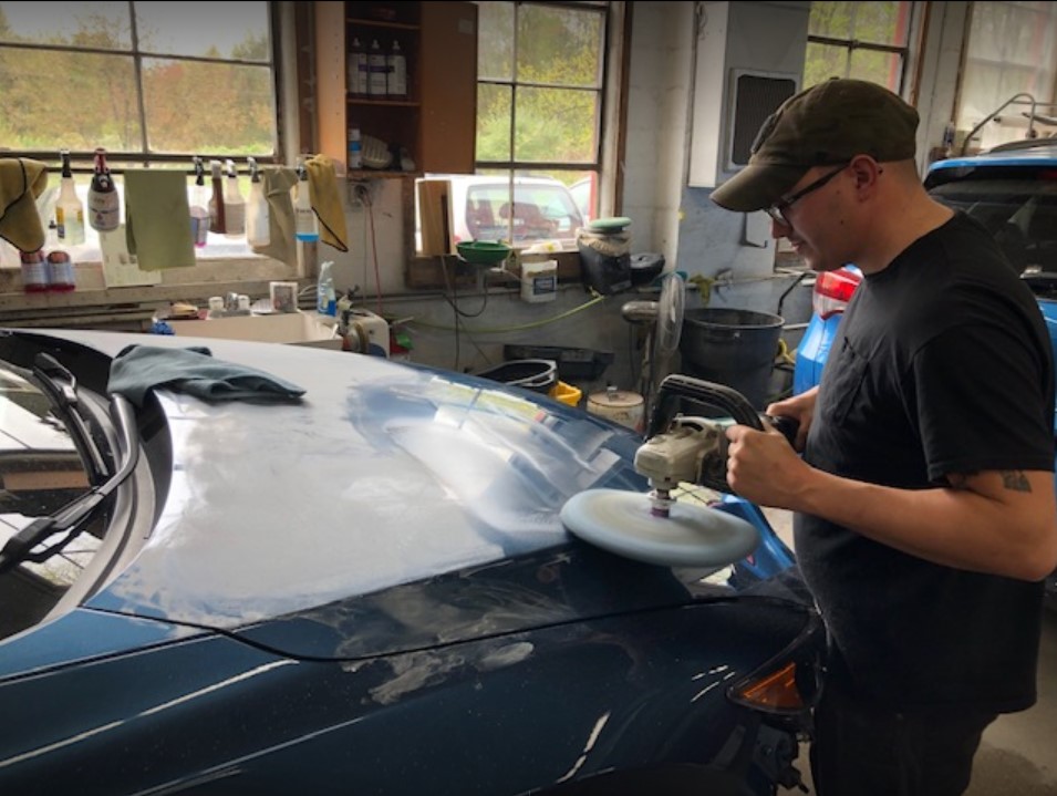A man wearing a baseball cap and glasses is buffing the hood of a blue car